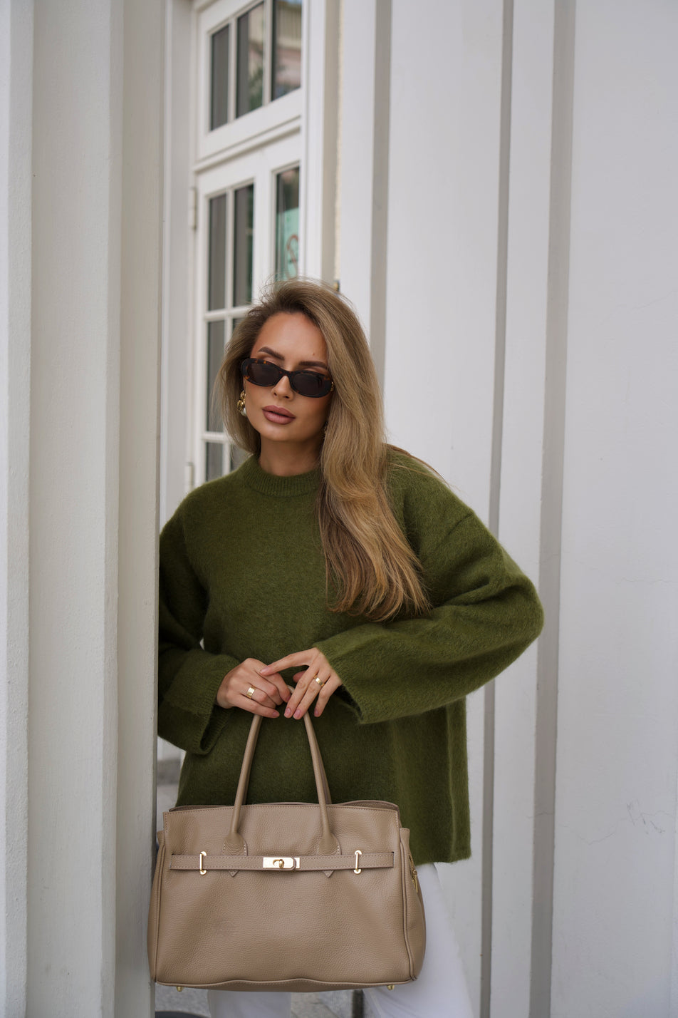 Lifestyle-Aufnahme der stehenden Frau, die die beige Henkeltasche stilvoll in der Hand trägt