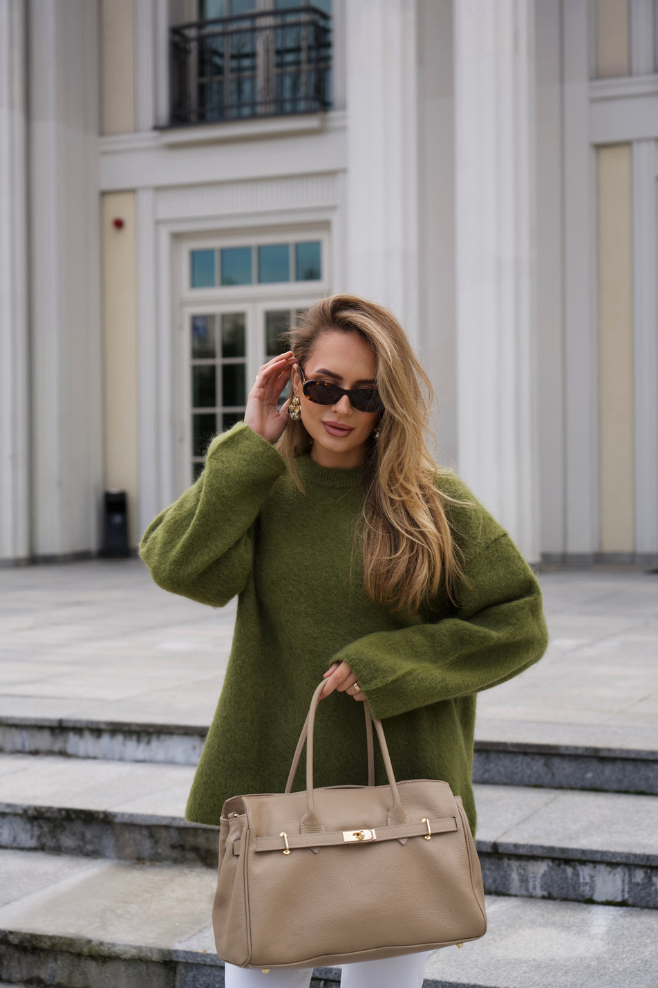 Model im olivgrünen Pullover präsentiert die beige Leder-Handtasche stehend auf Treppenstufen