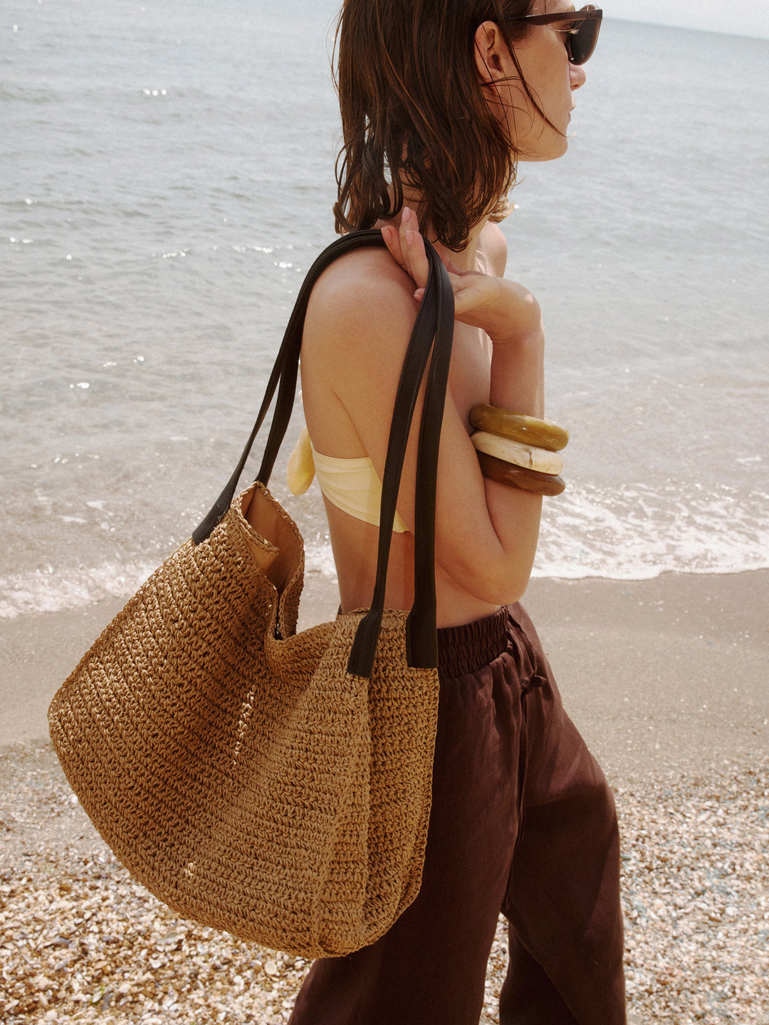 Beige Damen-Shopper-Tasche aus Stroh in künstlerischer Ansicht mit schwarzen Henkeln am Strand