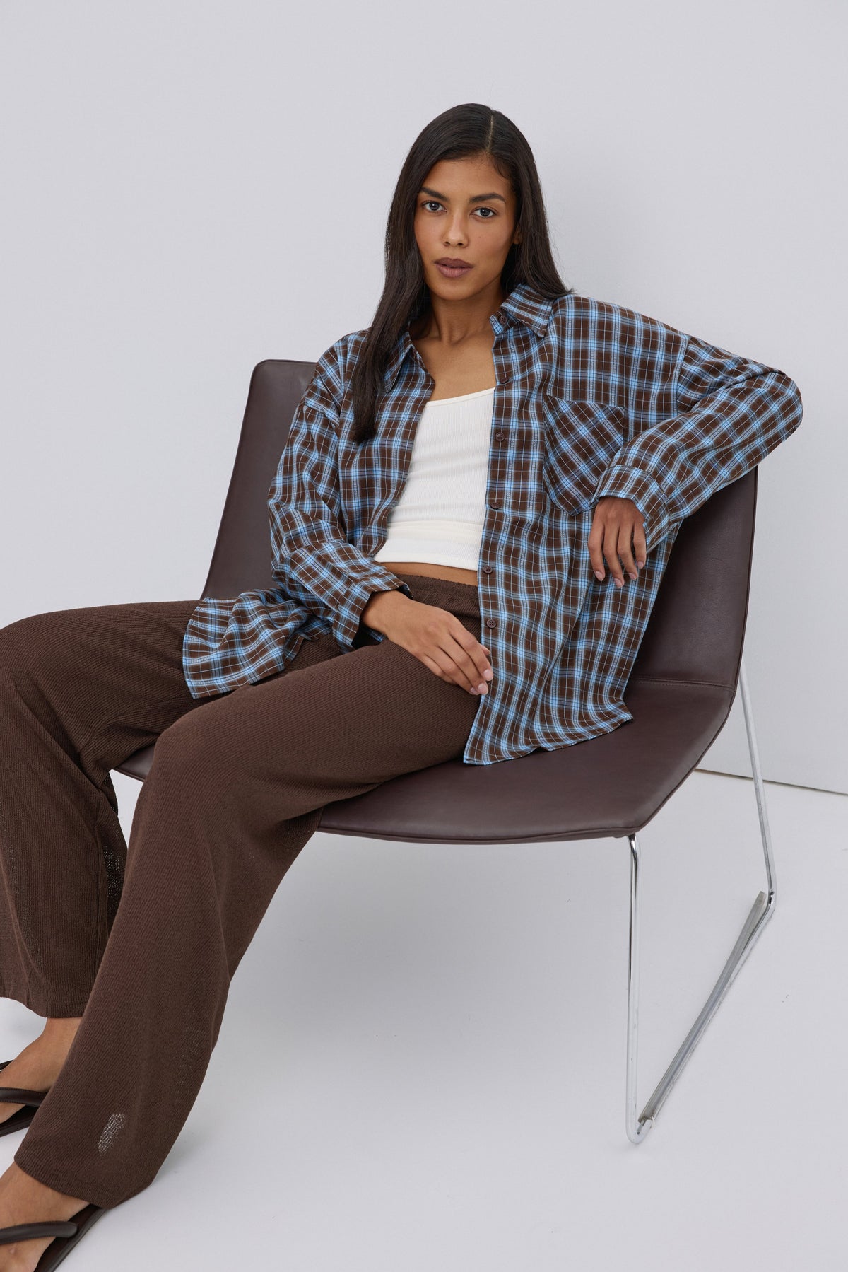 Model sitzt entspannt auf einem Sessel und präsentiert den lockeren Boxy-Fit der blau-braunen Hemdbluse