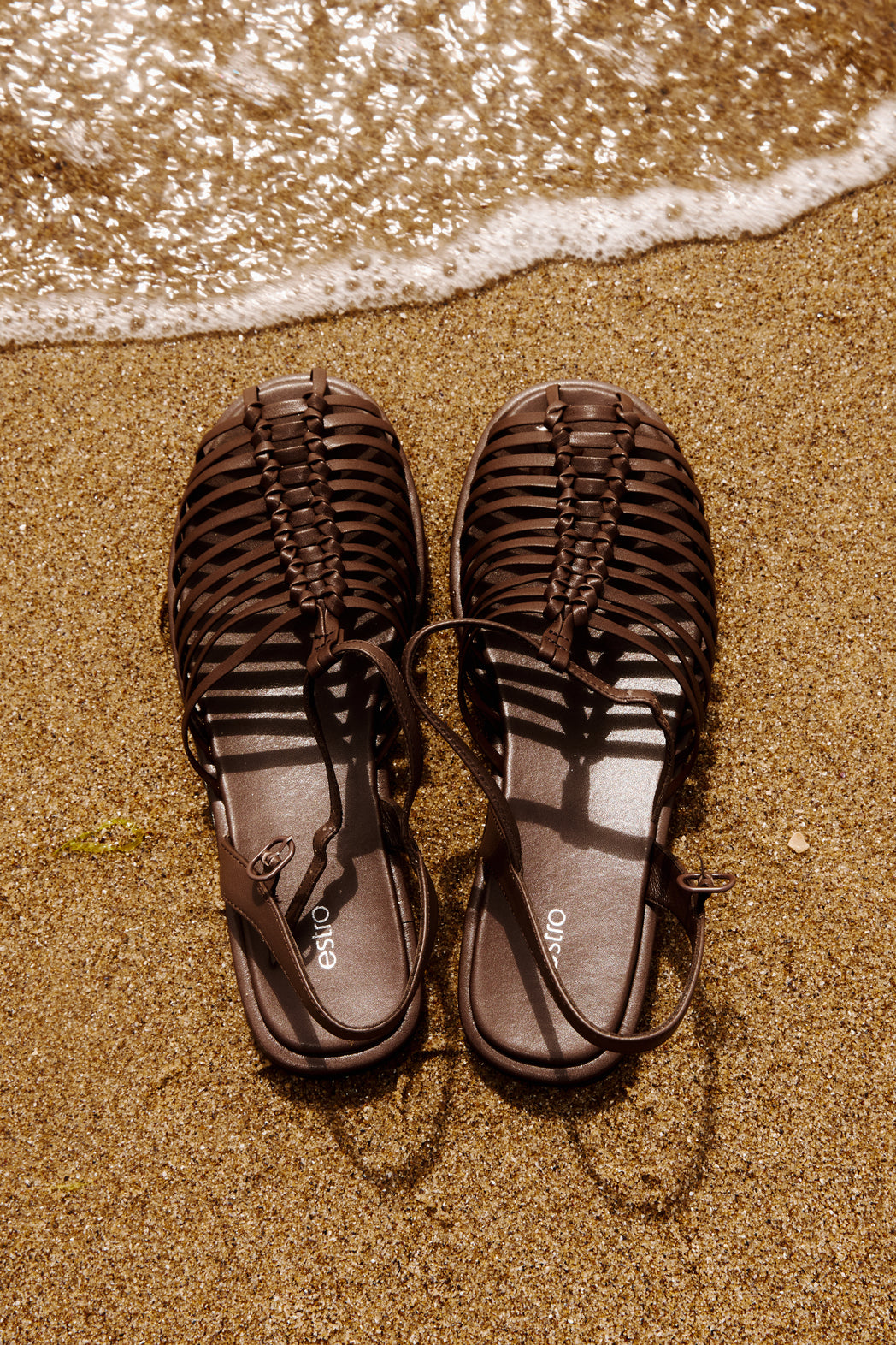 Draufsicht auf die dunkelbraunen Fisherman-Sandalen am Strand neben sanften Wellen