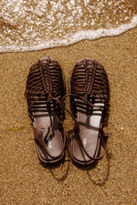 Draufsicht auf die dunkelbraunen Fisherman-Sandalen am Strand neben sanften Wellen