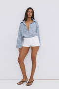 Model steht in einem sommerlichen Outfit mit blauem Hemd und weißen Shorts und präsentiert die dunkelbraunen Zehentrenner-Pantoletten