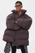 Model präsentiert die dunkelbraune Oversize-Pufferjacke mit Brille und Schal-Detail