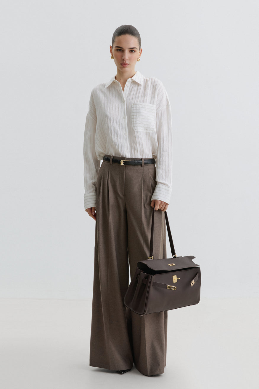 Model in weißer Bluse und weiter Stoffhose präsentiert die dunkelbraune Leder-Schultertasche 