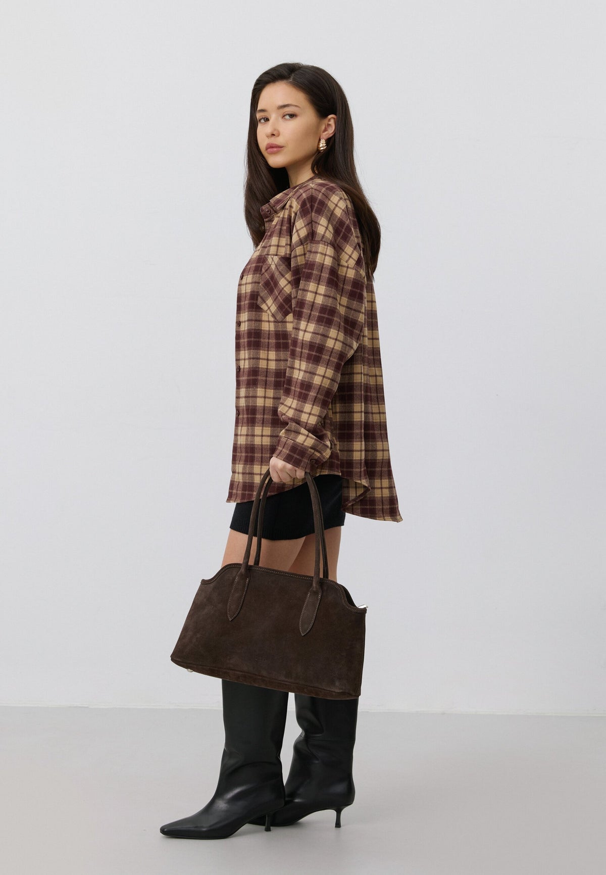 Lifestyle-Aufnahme einer Frau im karierten Hemd, die die dunkelbraune Velours-Schultertasche elegant in der Hand präsentiert