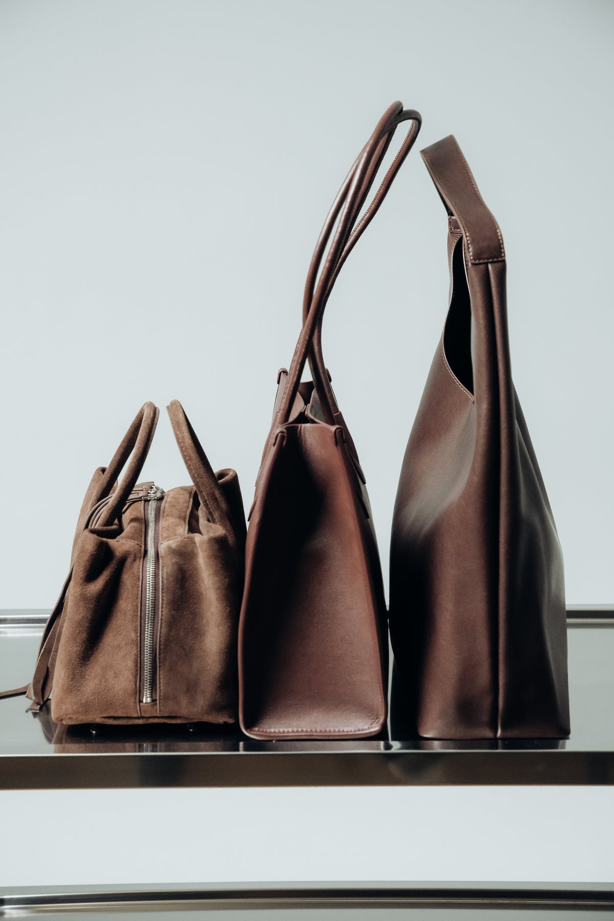 Ästhetischer Profil-Größenvergleich von drei dunkelbraunen Damen-Handtaschen aus Glatt- und Veloursleder auf einer Metalloberfläche