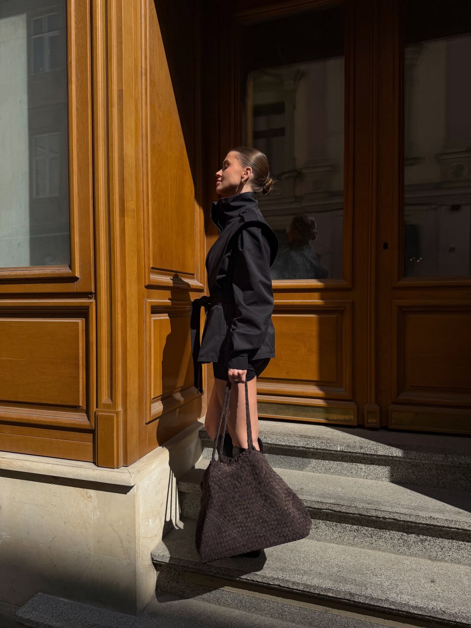 Ein Model auf einer sonnigen Treppe trägt die große dunkelbraune Shopper-Tasche aus geflochtenem Velours.