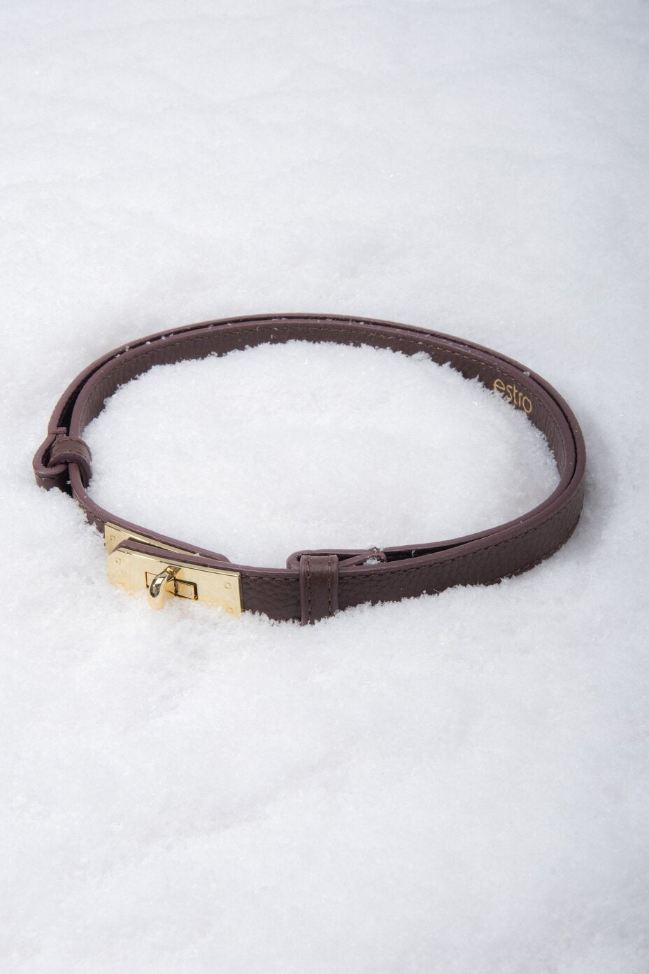 Kreative Produktinszenierung: Aufgerollter dunkelbrauner Damen-Gürtel aus Echtleder mit goldfarbenem Drehverschluss, liegend im weißen Schnee.
