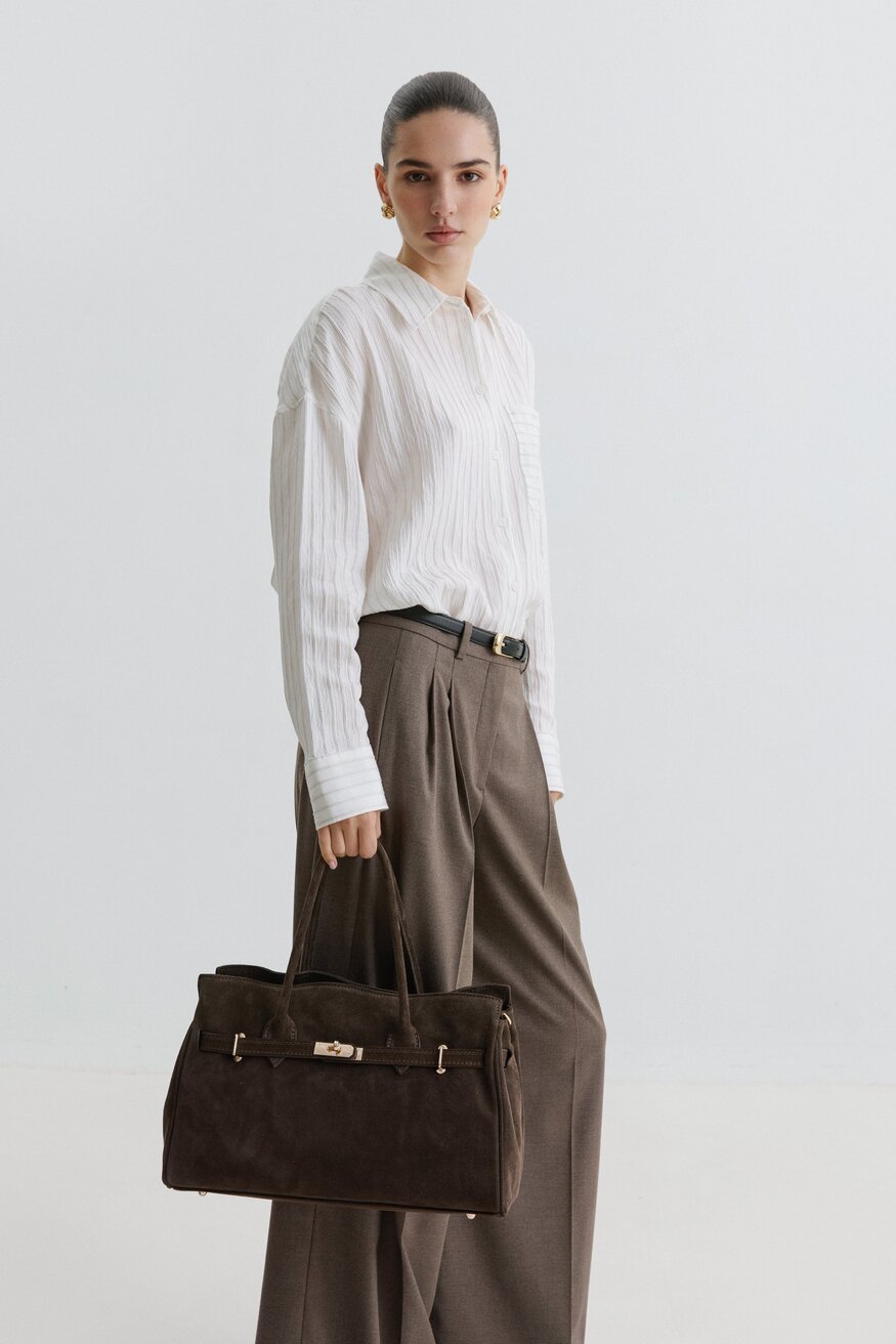Kampagnenbild eines Models in weißer Bluse und weiter Stoffhose, das die dunkelbraune Velours-Handtasche trägt