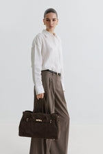 Kampagnenbild eines Models in weißer Bluse und weiter Stoffhose, das die dunkelbraune Velours-Handtasche trägt