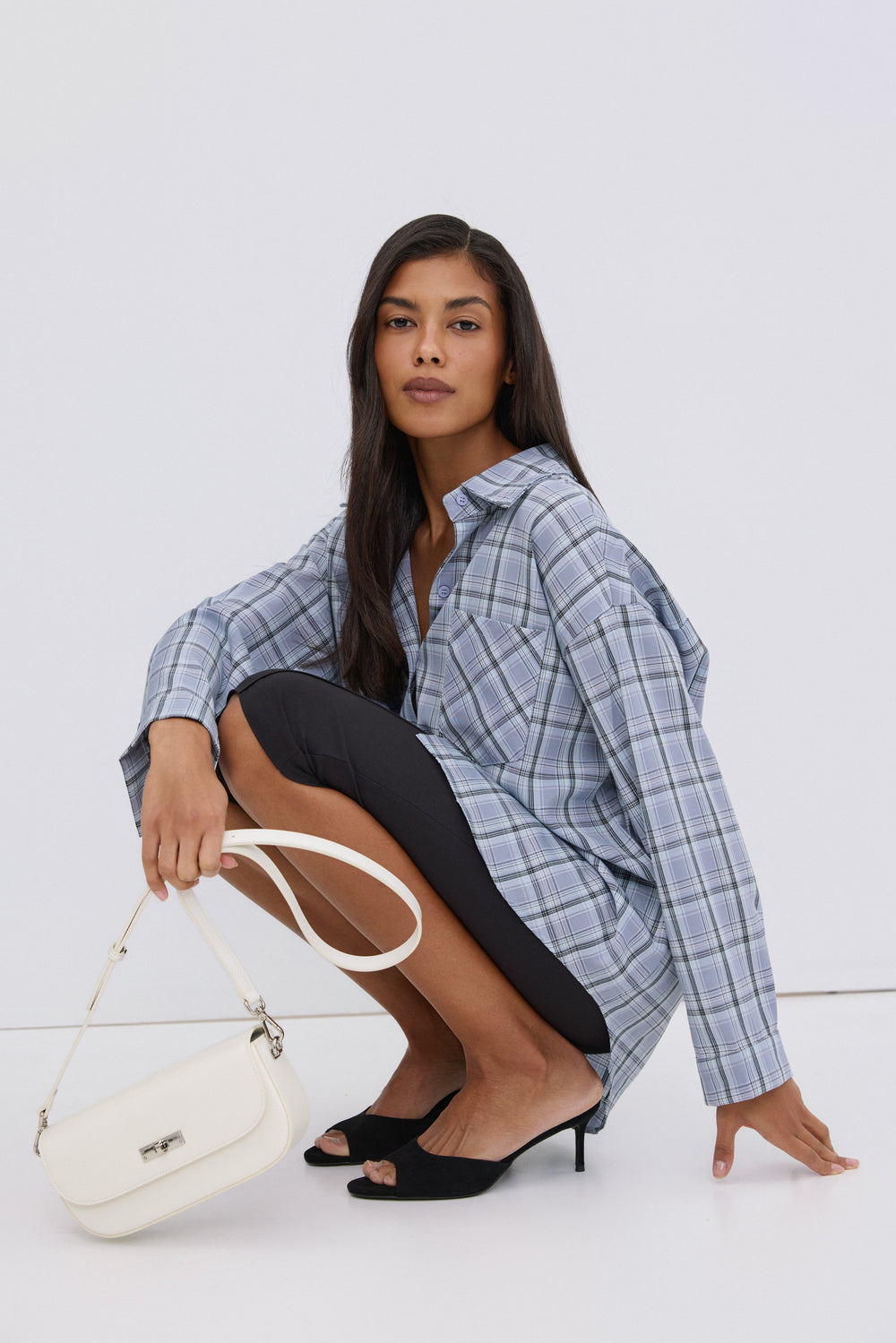 Model hockend in der hellblau karierten Oversize-Hemdbluse, kombiniert mit einer weißen Estro-Handtasche