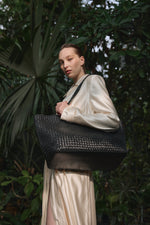 Ein Model in einer üppigen grünen Kulisse trägt die große schwarze Shopper-Tasche aus geflochtenem Leder.