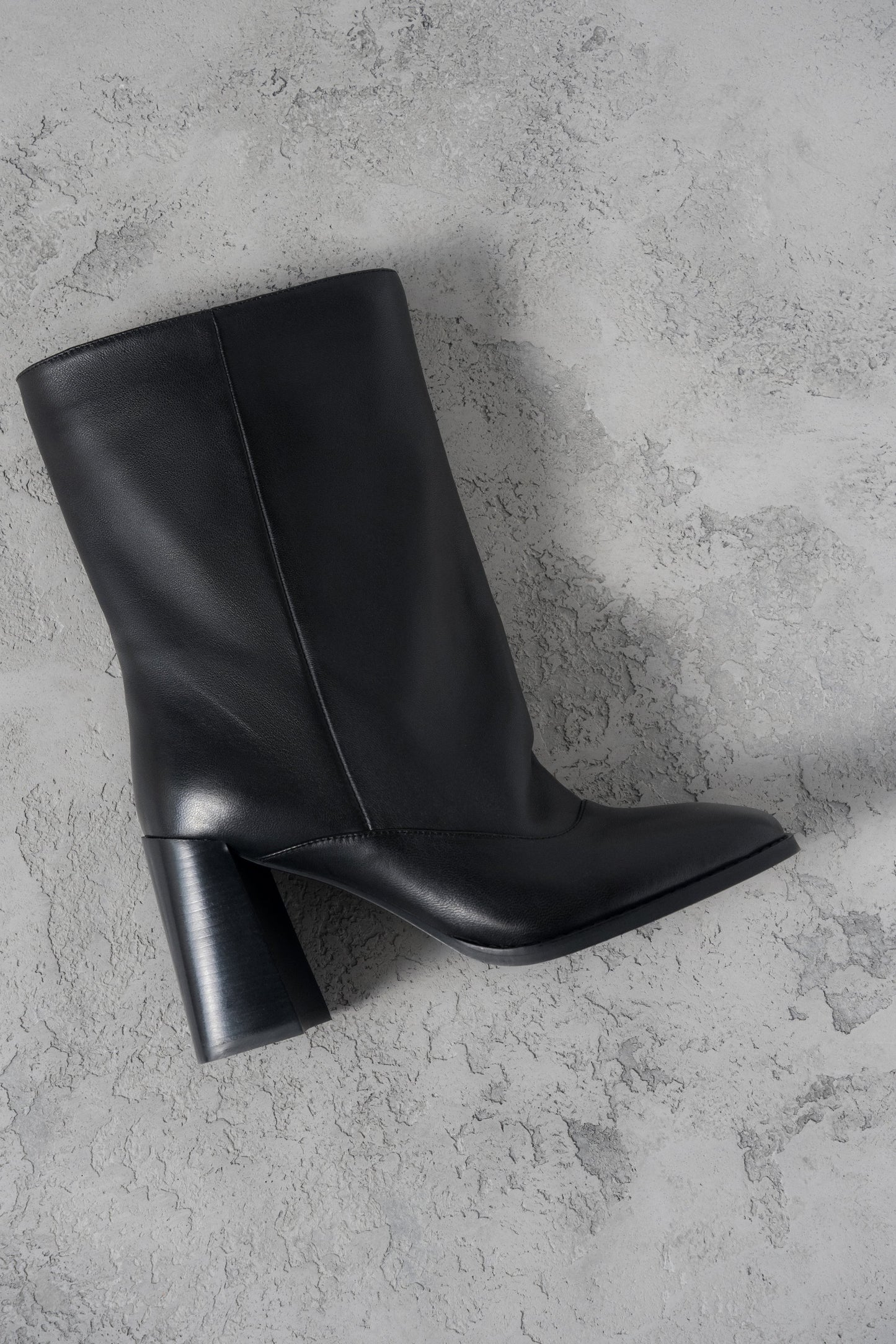Atmosphärische Aufnahme der schwarzen Leder-Stiefelette mit weitem Schaft und Karree-Spitze auf grauem Untergrund
