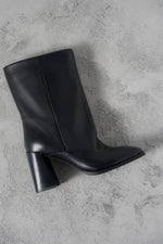 Atmosphärische Aufnahme der schwarzen Leder-Stiefelette mit weitem Schaft und Karree-Spitze auf grauem Untergrund