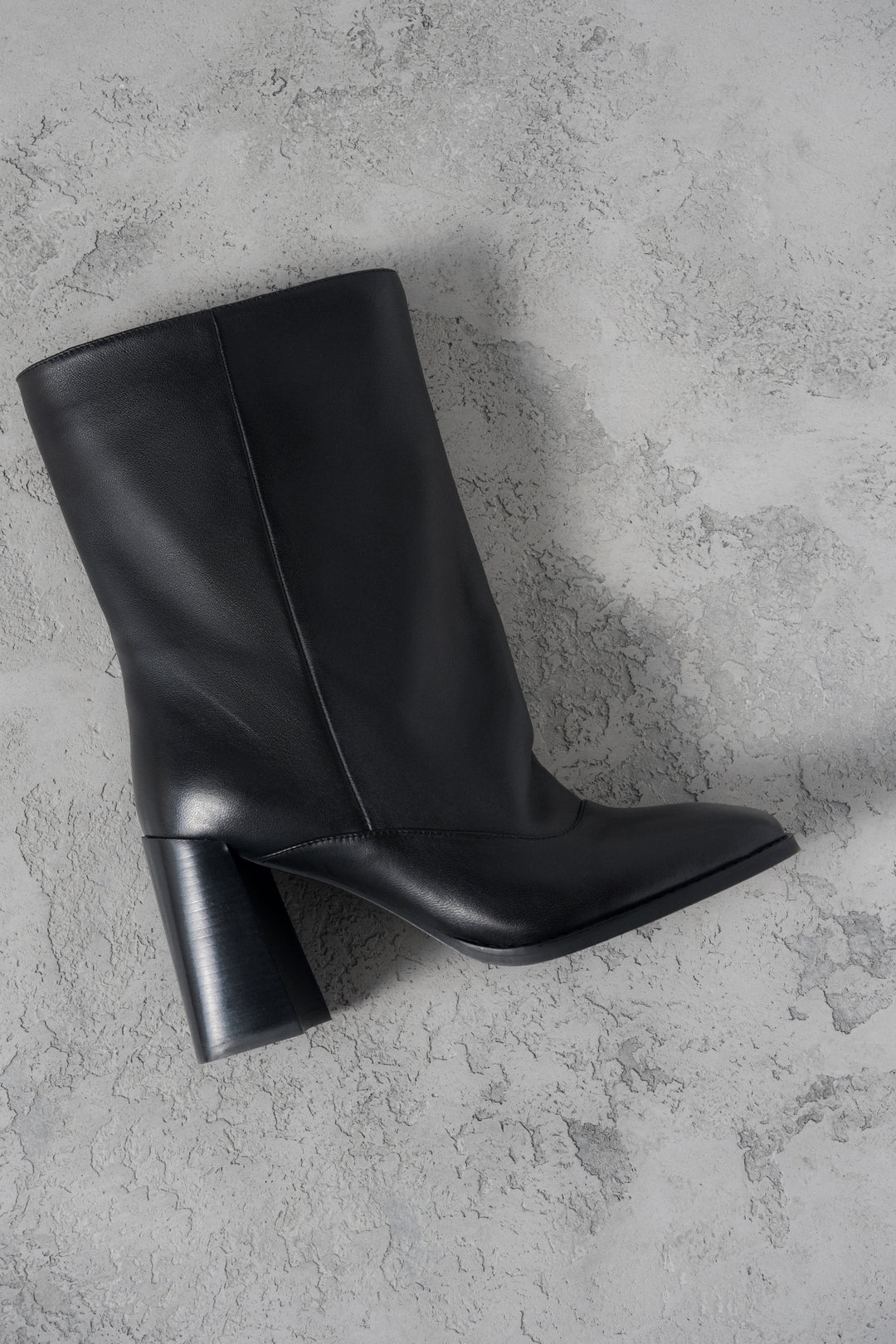 Atmosphärische Aufnahme der schwarzen Leder-Stiefelette mit weitem Schaft und Karree-Spitze auf grauem Untergrund